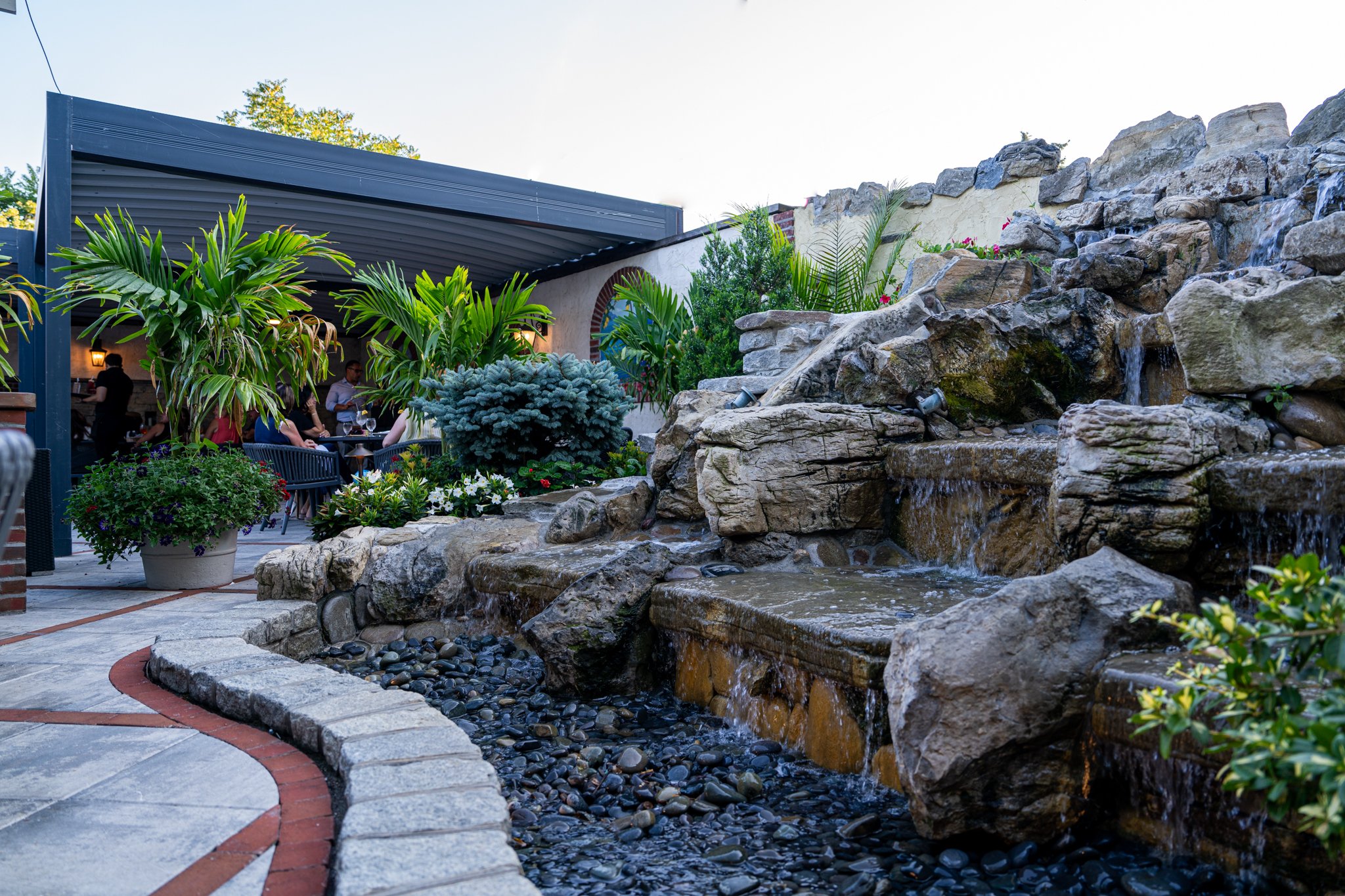 Outdoor patio with a waterfall feature, potted plants, and a dining area with people sitting under a metal roof.