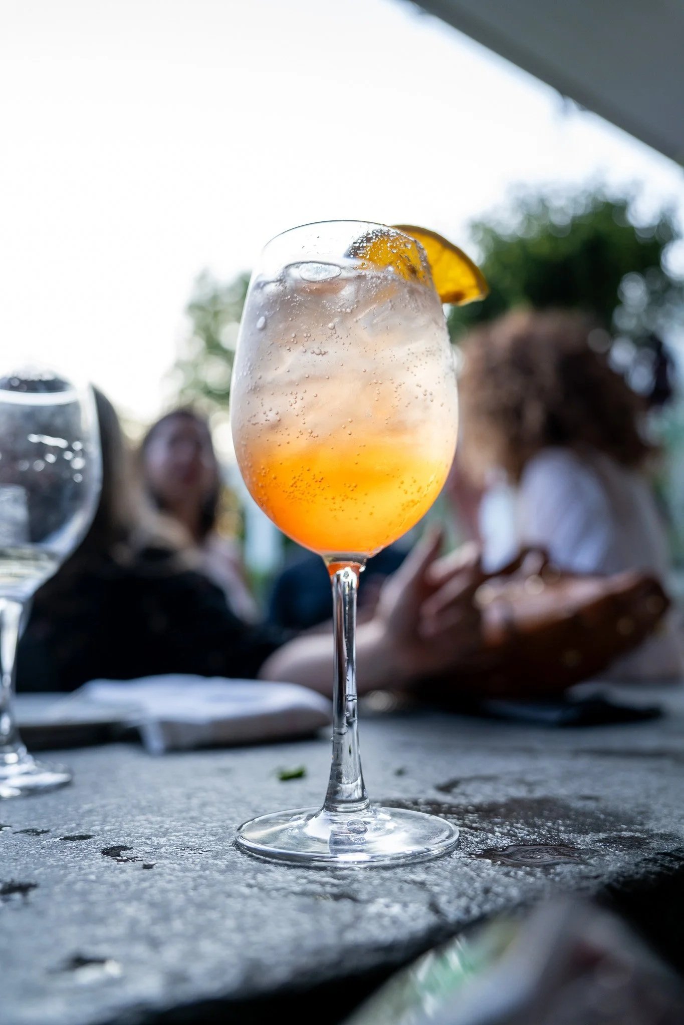 A glass of cocktail with orange slice garnish, bubbling and ice, outdoors with blurred people in background.