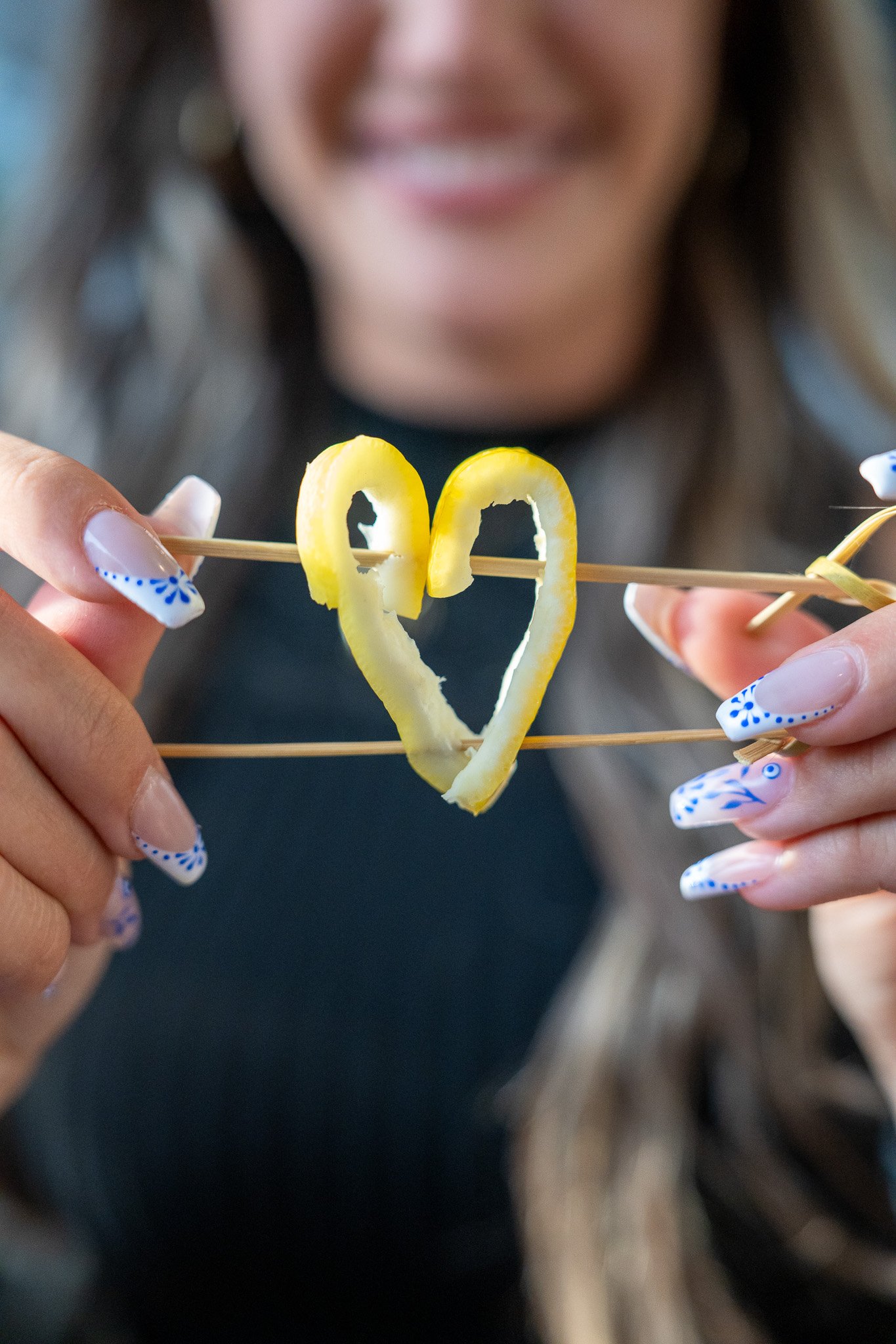A person holding two skewers of curled citrus peel formed into a heart shape.