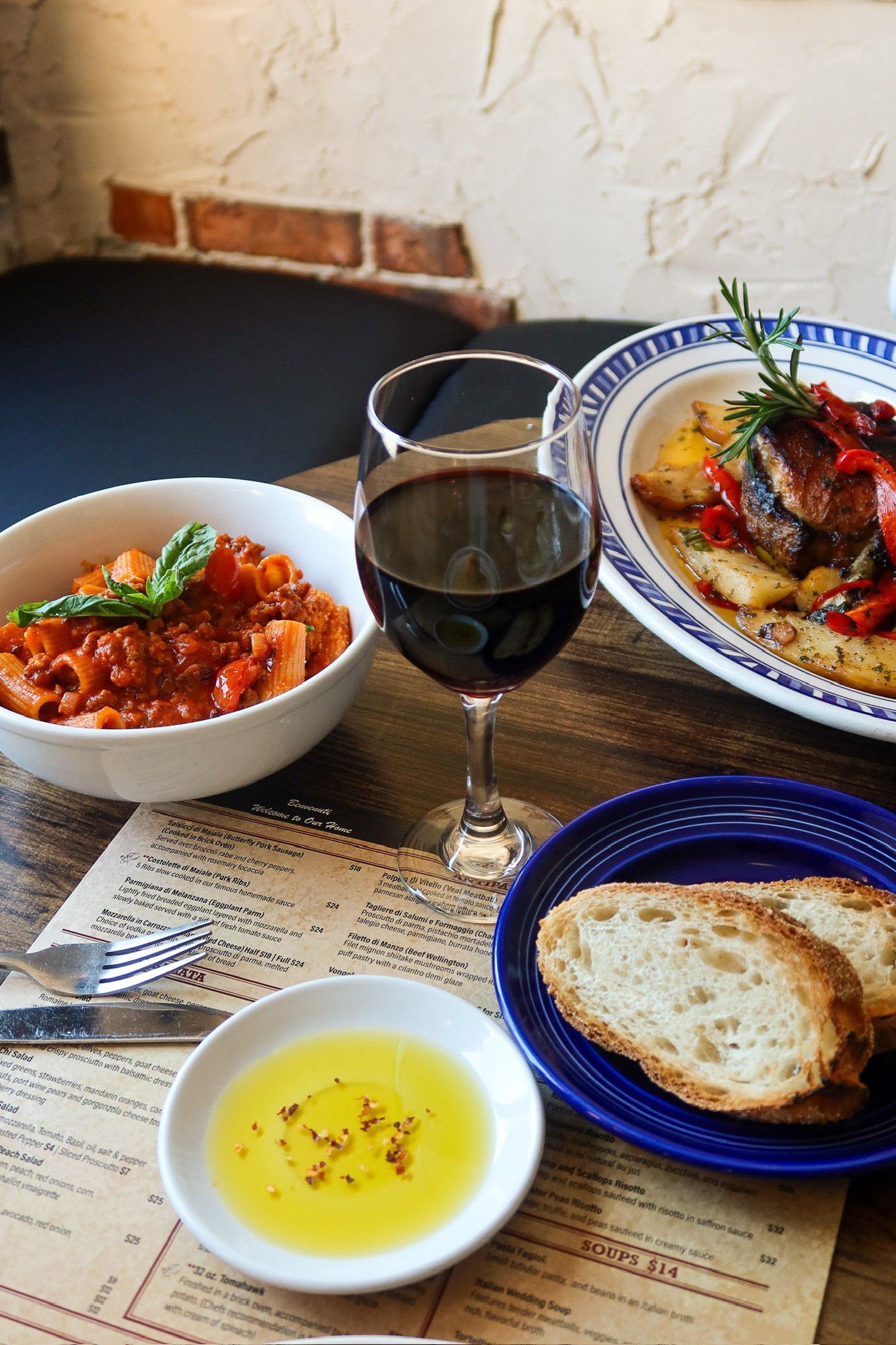 A glass of red wine, a bowl of pasta with tomato sauce and basil, a plate with roasted meat and potatoes, slices of bread, and a small dish of olive oil with seasoning on a wooden table in a restaurant setting.