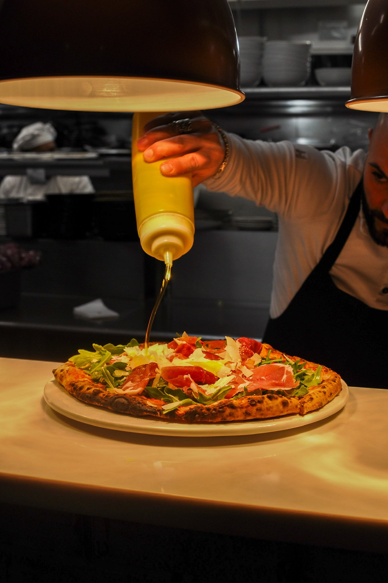 A chef drizzling yellow sauce on a pizza topped with vegetables and cured meat in a restaurant kitchen.