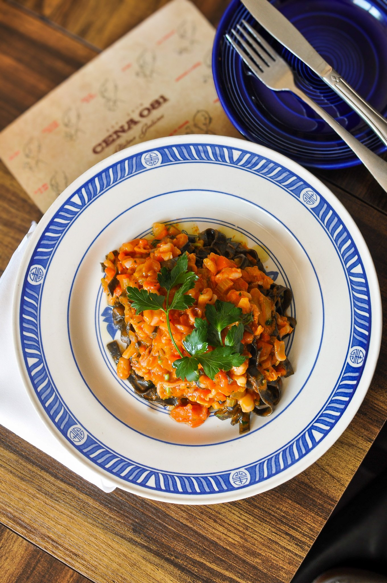 A plate of vegetable pasta with tomato sauce garnished with parsley, served on a white dish with blue patterns, placed on a wooden table.