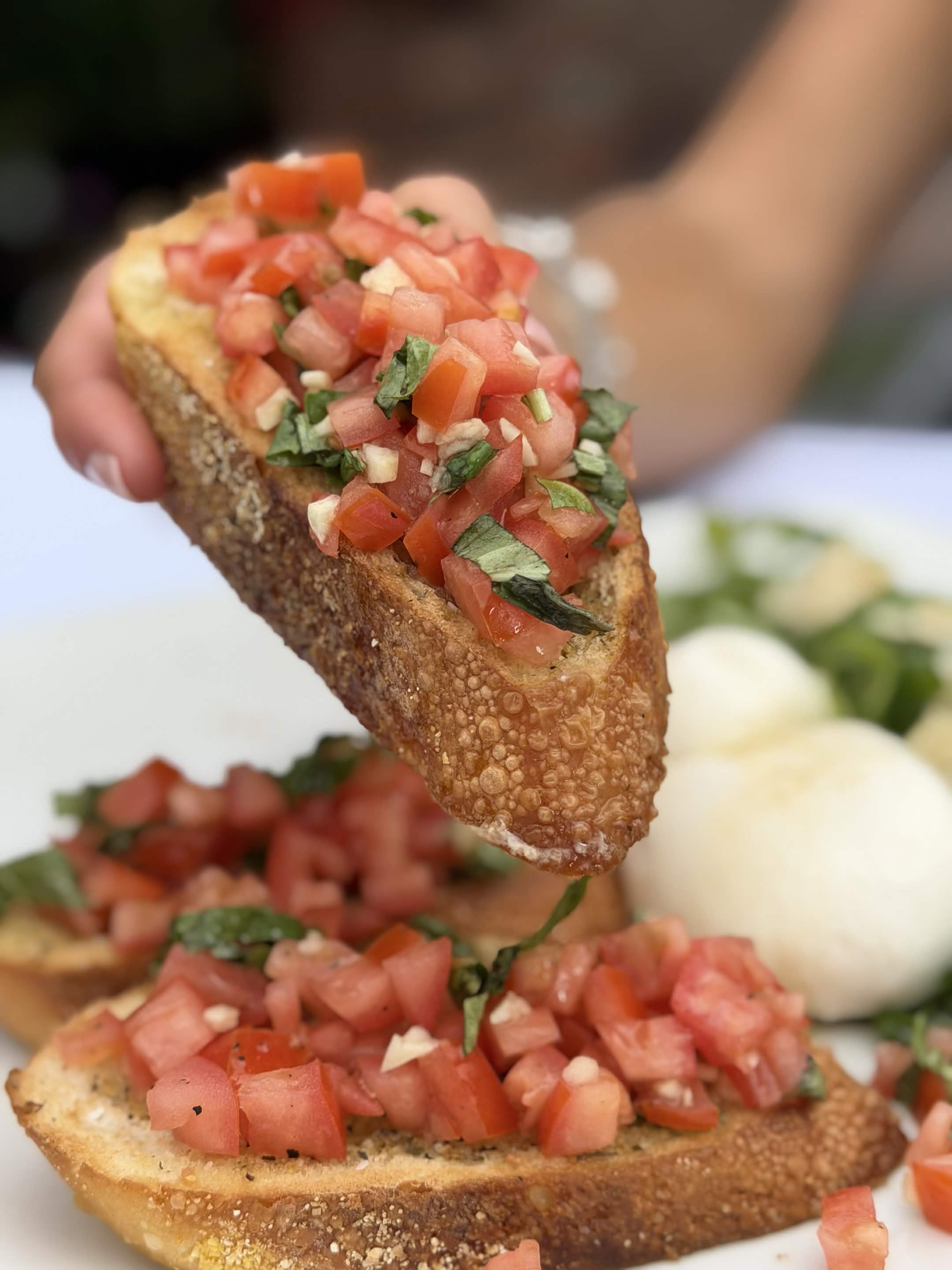 A hand lifting a slice of crusty bruschetta crostini piled with diced tomato, basil and garlic, with burrata and more crostini on the plate below.