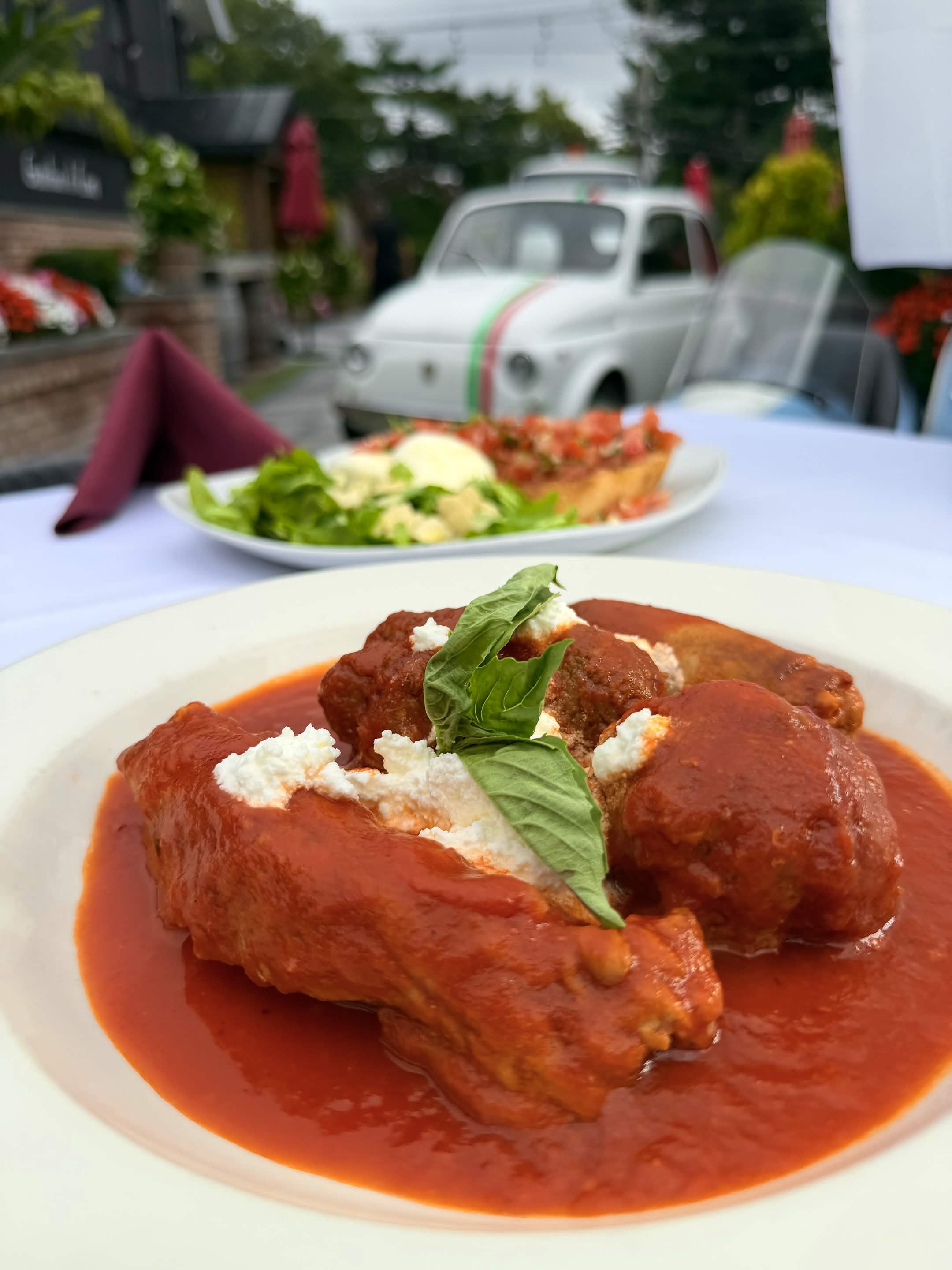 A Sunday meat sampler of meatball, braciola and pork ribs in marinara topped with fresh ricotta and basil, with a bruschetta and burrata plate behind it on the patio.