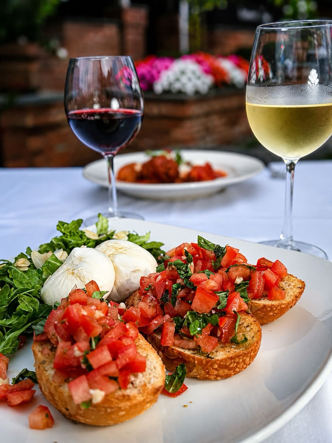 Toasted crostini topped with tomato-basil bruschetta beside a ball of burrata and dressed arugula, with glasses of red and white wine on a white tablecloth.