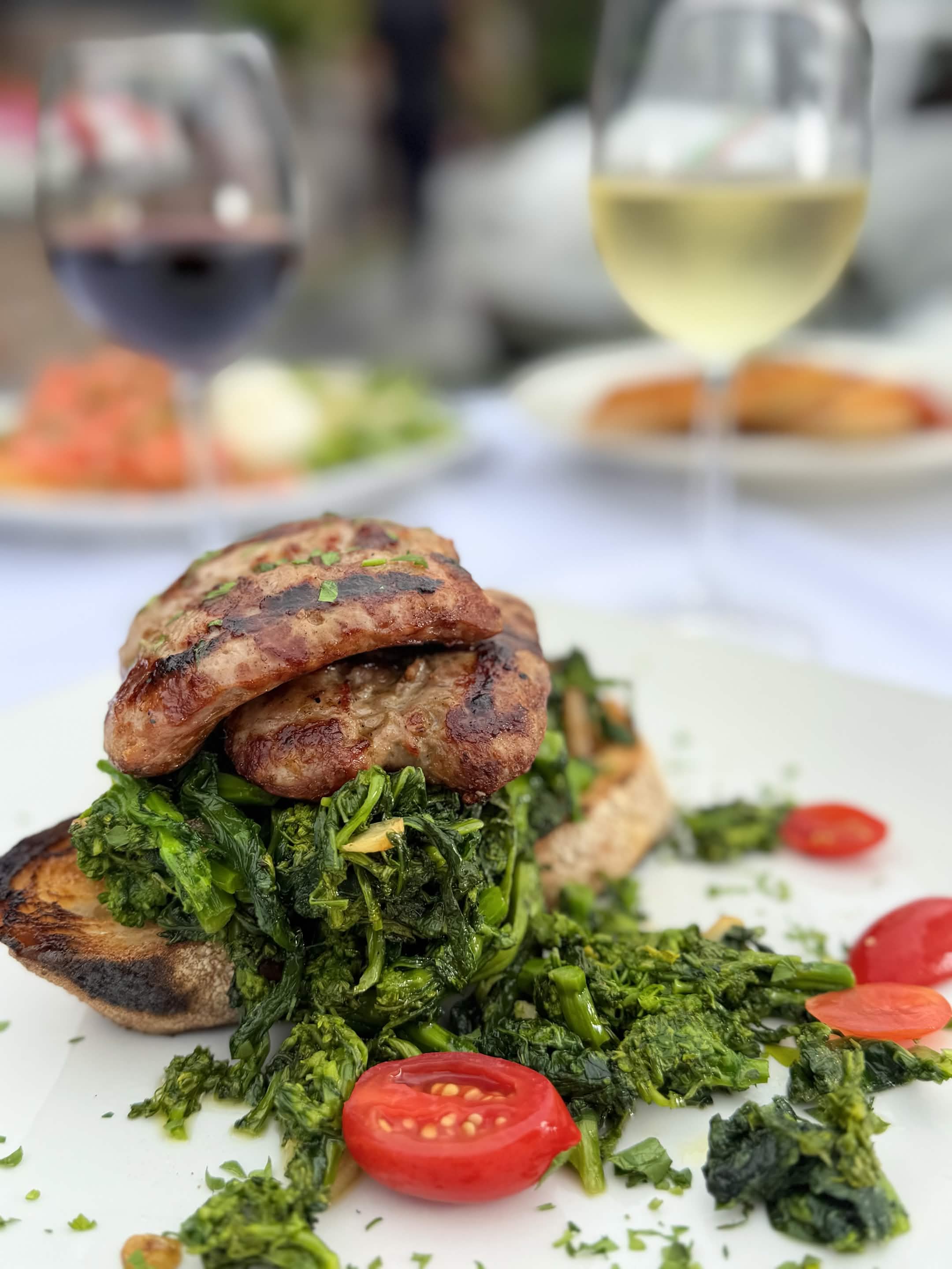 Grilled Italian sausage served over sautéed broccoli rabe on toasted garlic bread, garnished with halved cherry tomatoes.